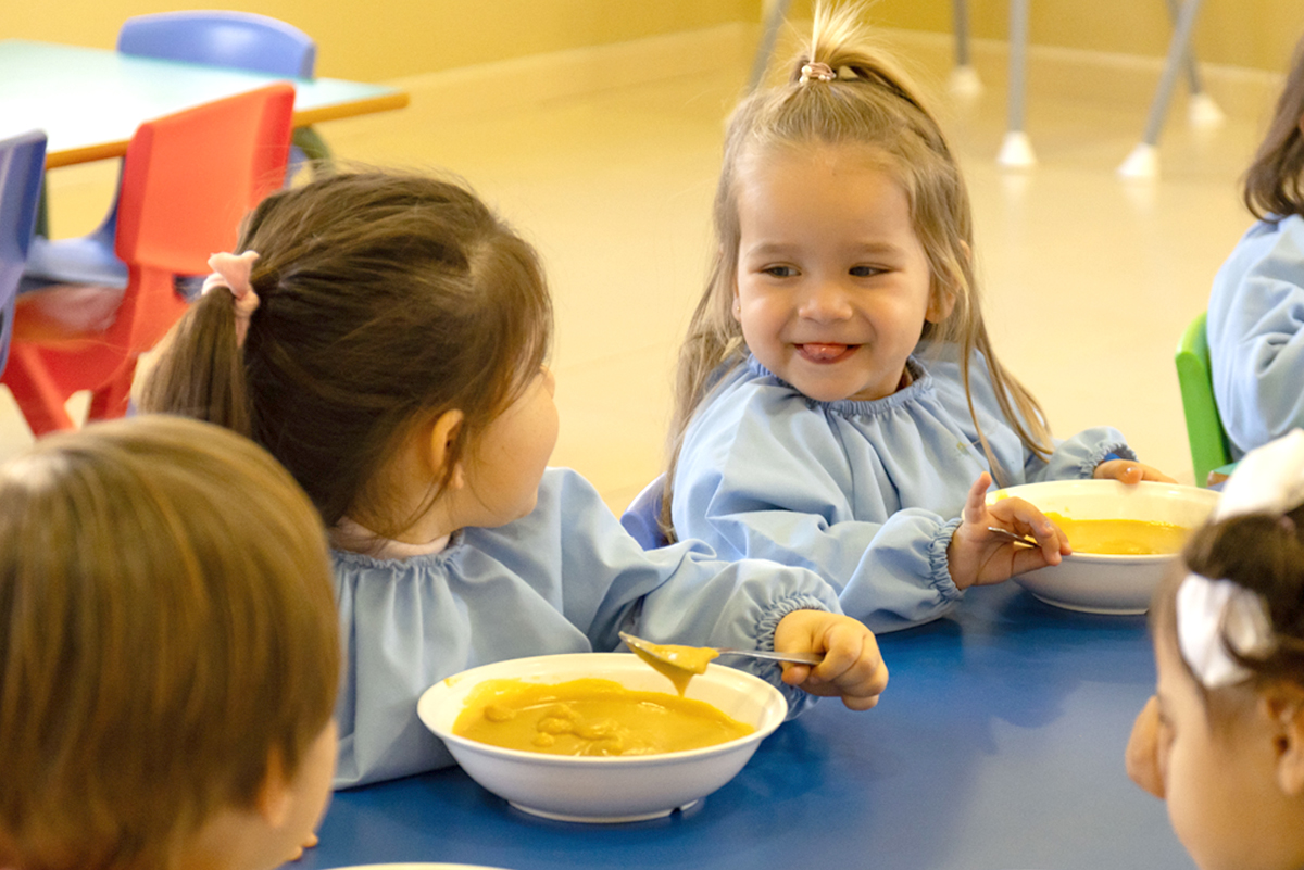 Dos niñas comiendo puré y mirándose una a la otra mientras una de ellas sonríe y saca la lengua.